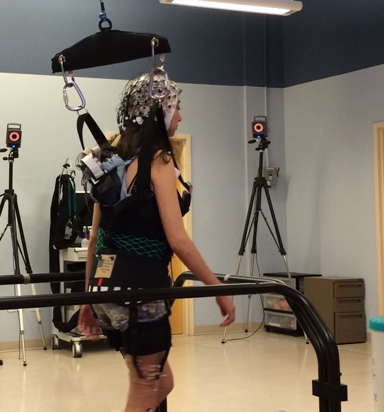 A person in a harness walking on a treadmill in a rehabilitation center, with monitoring equipment and cameras set up around.