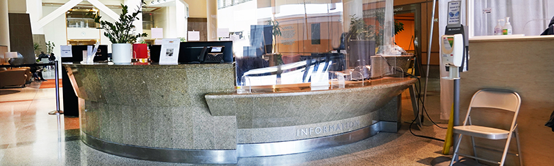 The information desk in the main lobby at the NIH Clinical Center, with a clear protective barrier, office equipment, and a hand sanitizer station nearby.