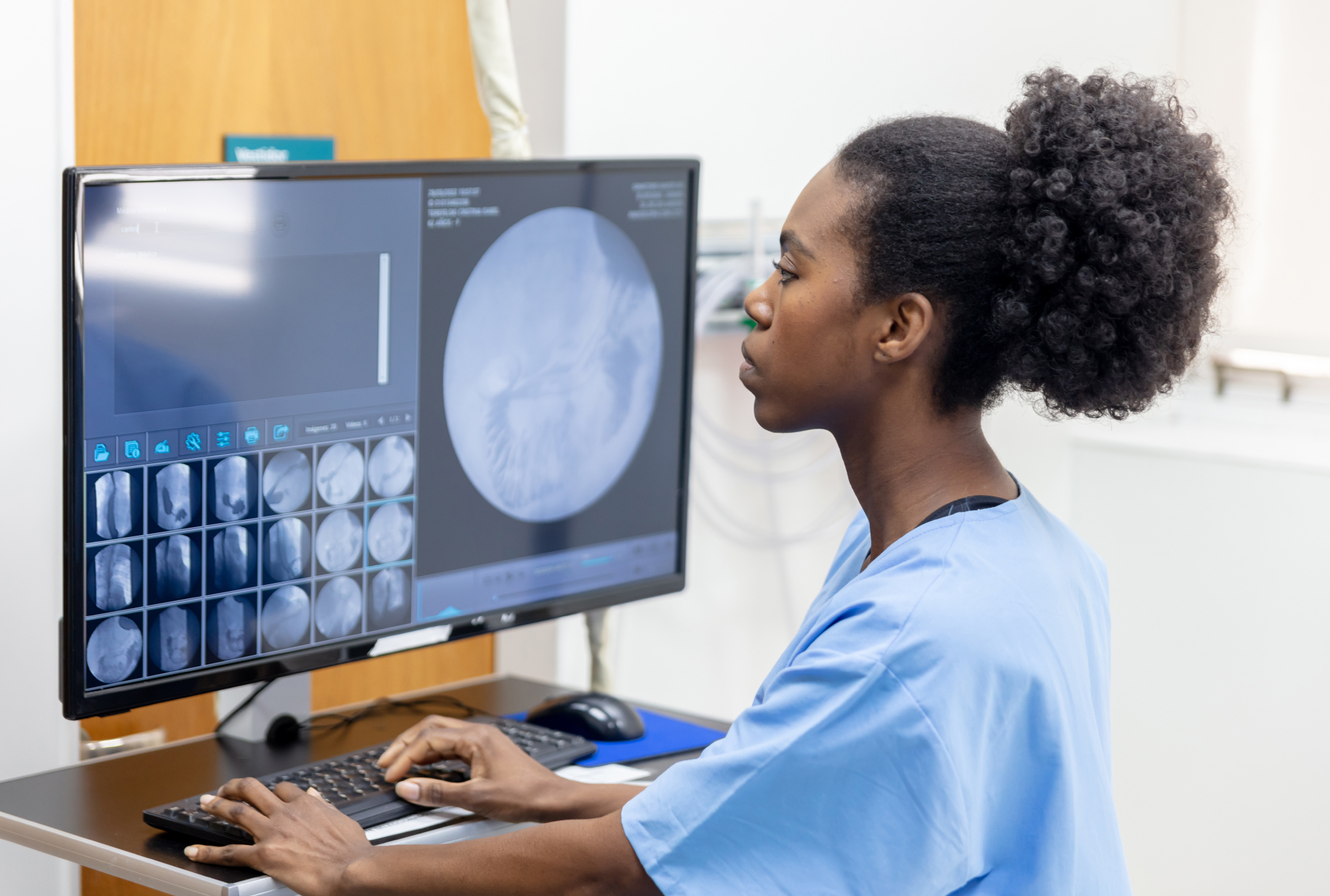 A radiology technician in scrubs working at a computer with medical imaging displayed on the monitor.