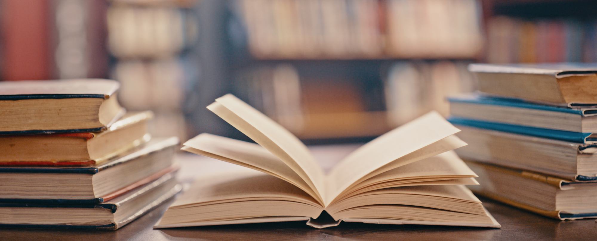 An open book on a table with stacks of books in the background.