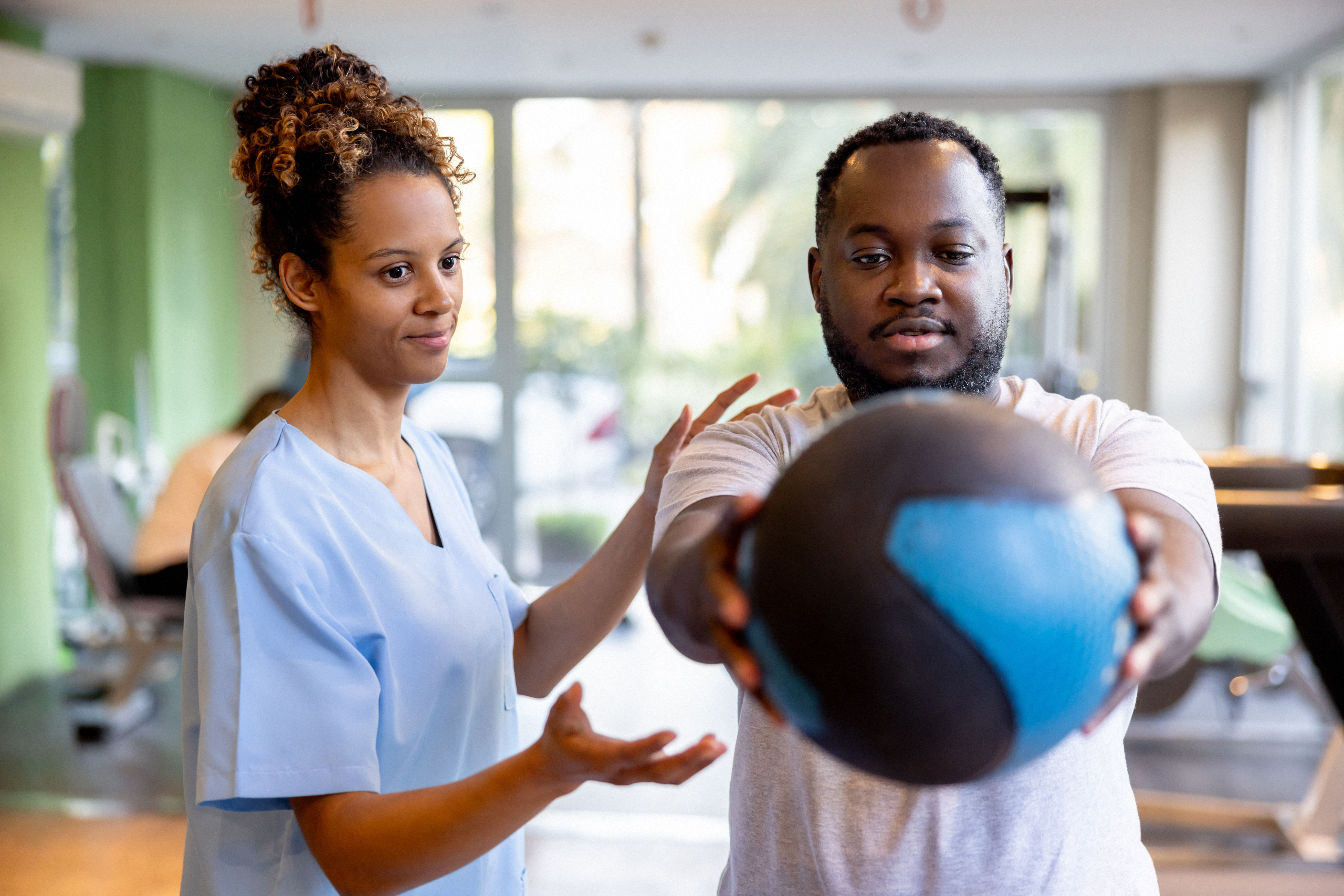 A healthcare professional guides a patient during physical therapy as he holds a medicine ball in a gym-like setting.